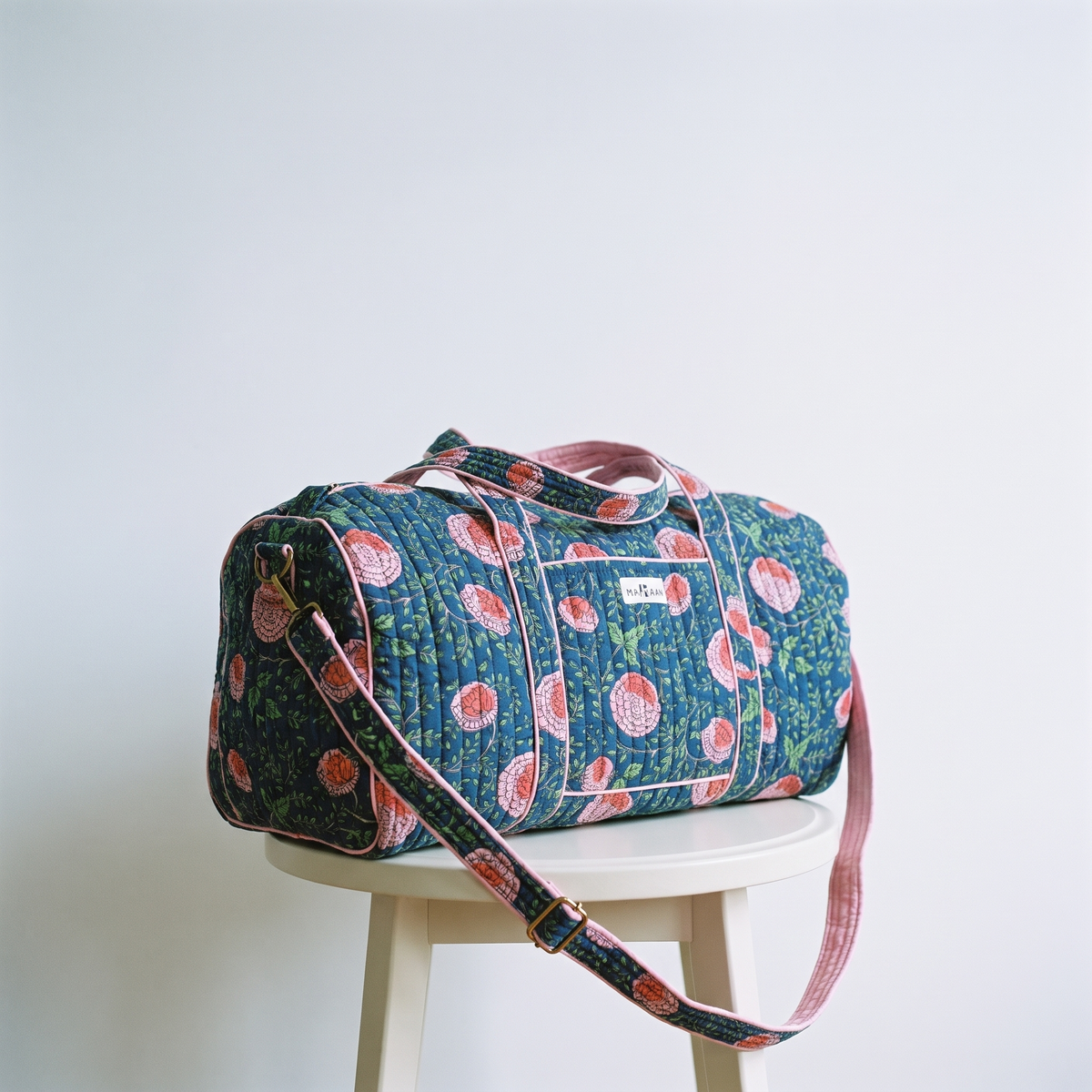 Sac week-end Kusum posé seul sur un tabouret clair, bandoulière retombant en courbe