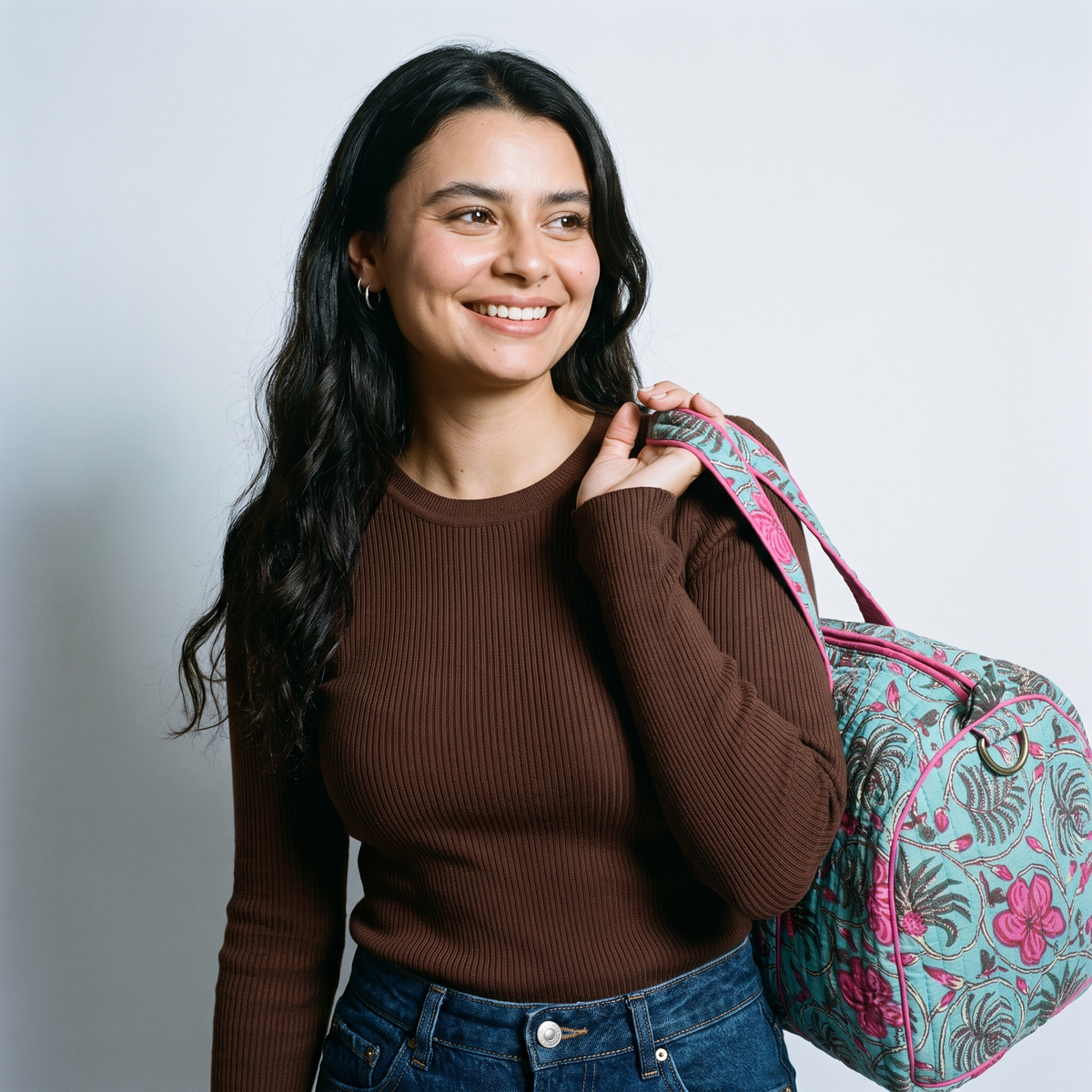 Femme souriante portant le sac week-end Nilofar à l’épaule par ses anses, blockprint turquoise et fleurs fuchsia, sur un pull chocolat